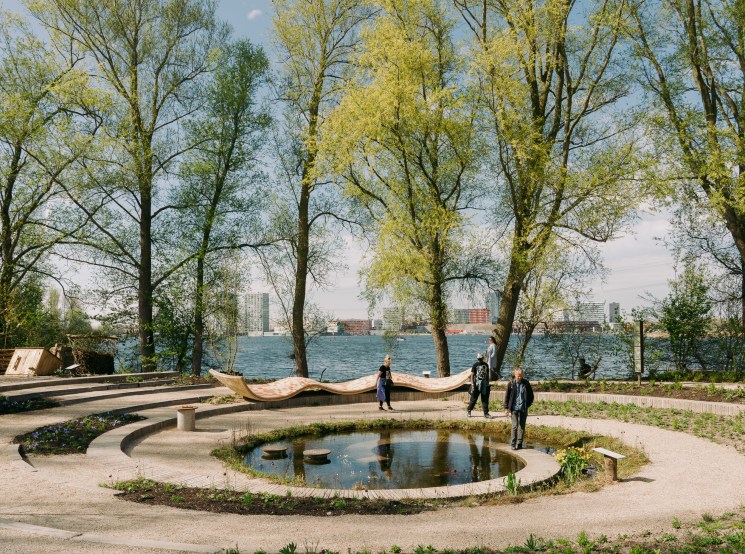 Community garden with circular pond, wooden benches, and people walking along waterfront with trees and city skyline