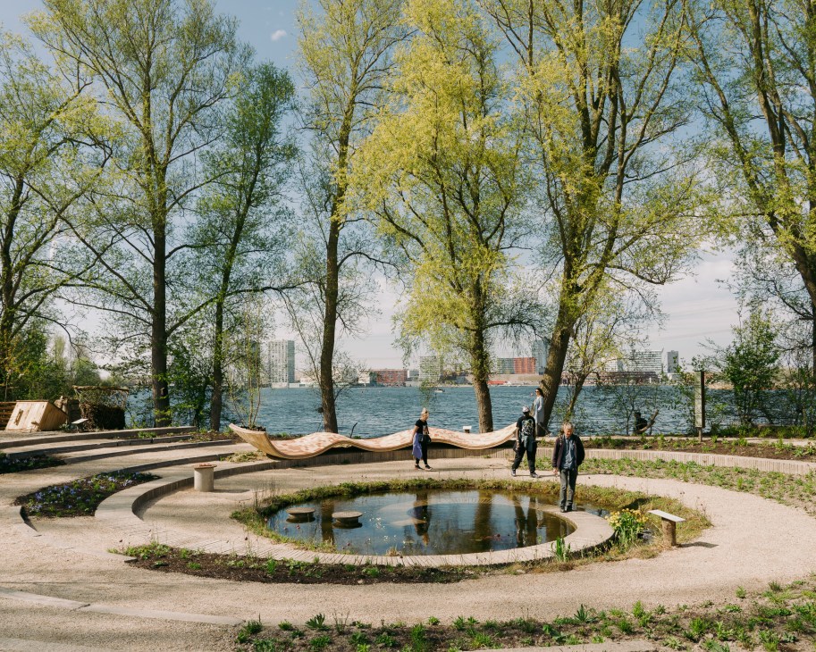 Community garden with circular pond, wooden benches, and people walking along waterfront with trees and city skyline