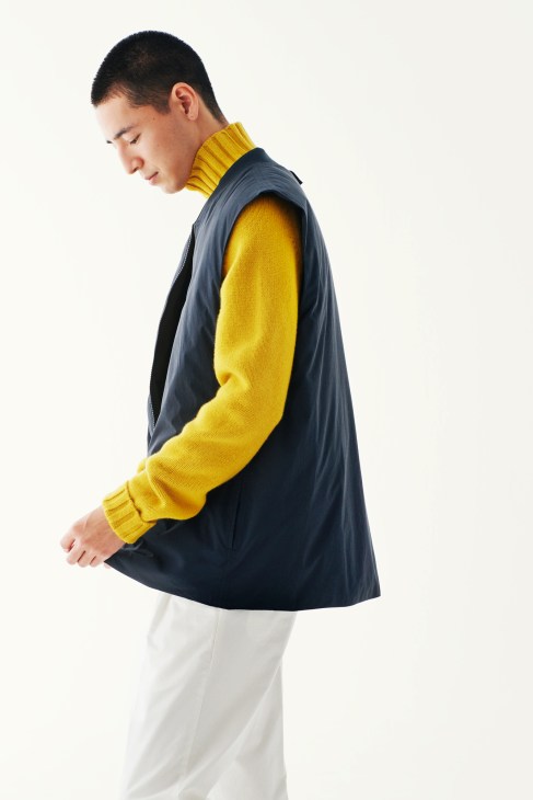 Man in profile wearing yellow turtleneck, navy vest, and light trousers against white background.