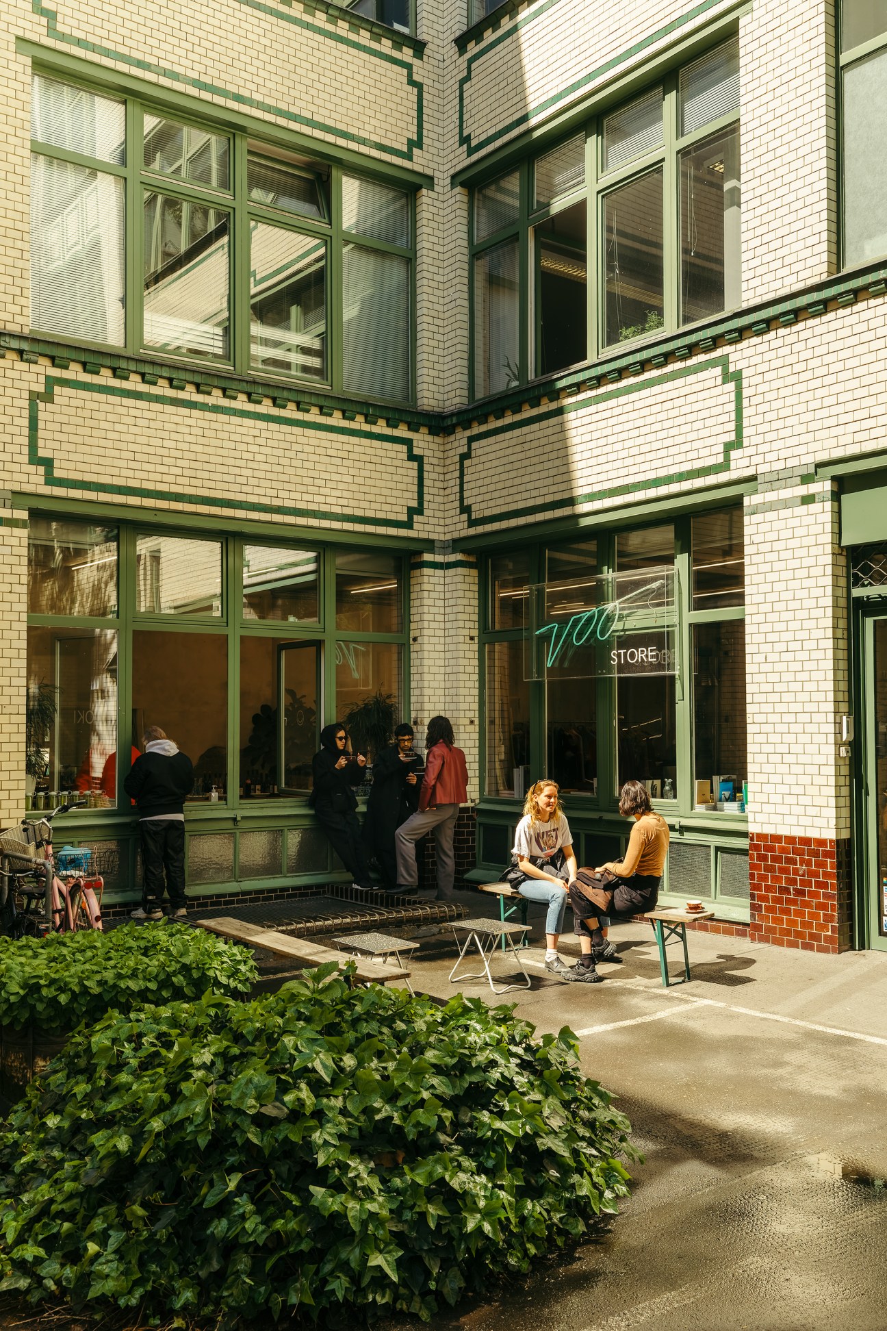 Customers relax in a sunny brick courtyard outside Voo Store with green-framed windows and potted plants.