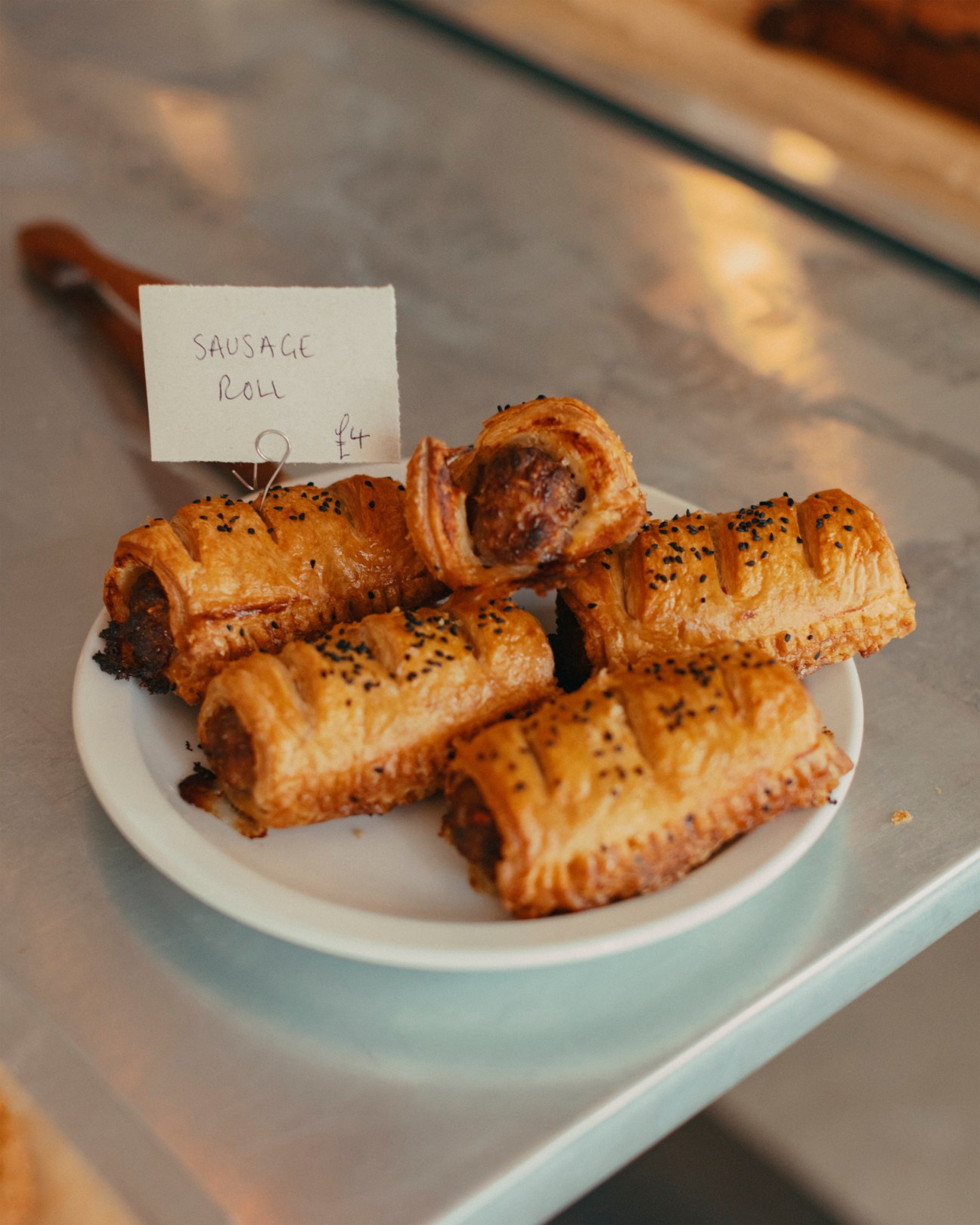 Golden puff pastry sausage rolls topped with black seeds displayed on white plate with price tag showing £4