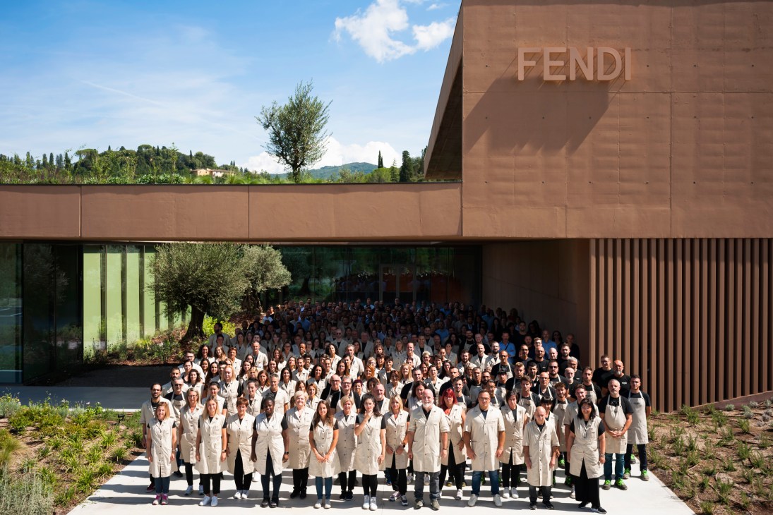 Large group of Fendi artisans in white coats gathered outside modern brown stone factory building in Tuscany