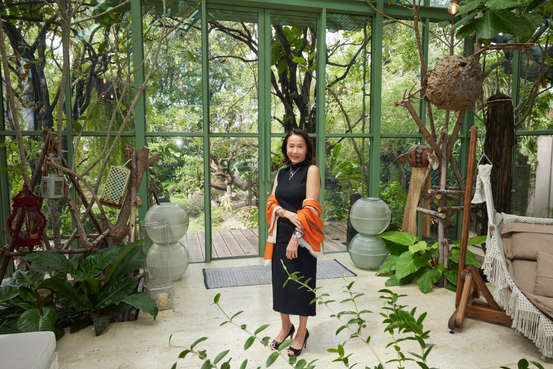 Woman in black dress with orange cardigan standing in glass-walled greenhouse space with plants and decorative objects
