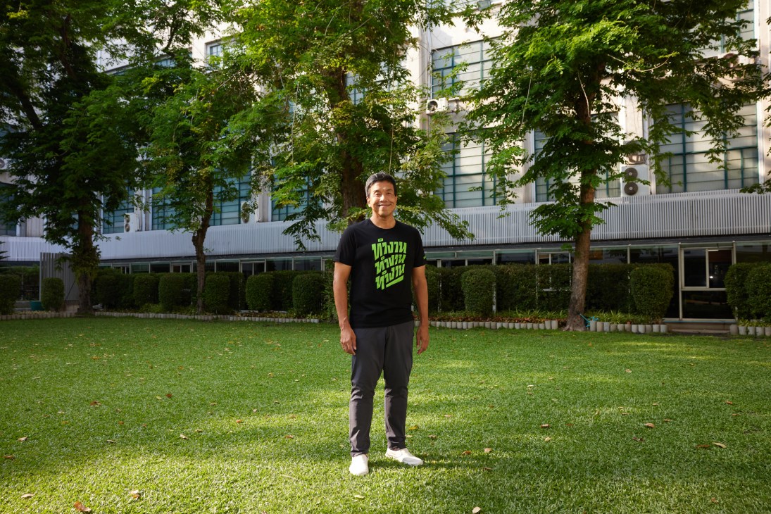 Chadchart Sittipunt standing on grass in front of a modern building wearing a black t-shirt with green text