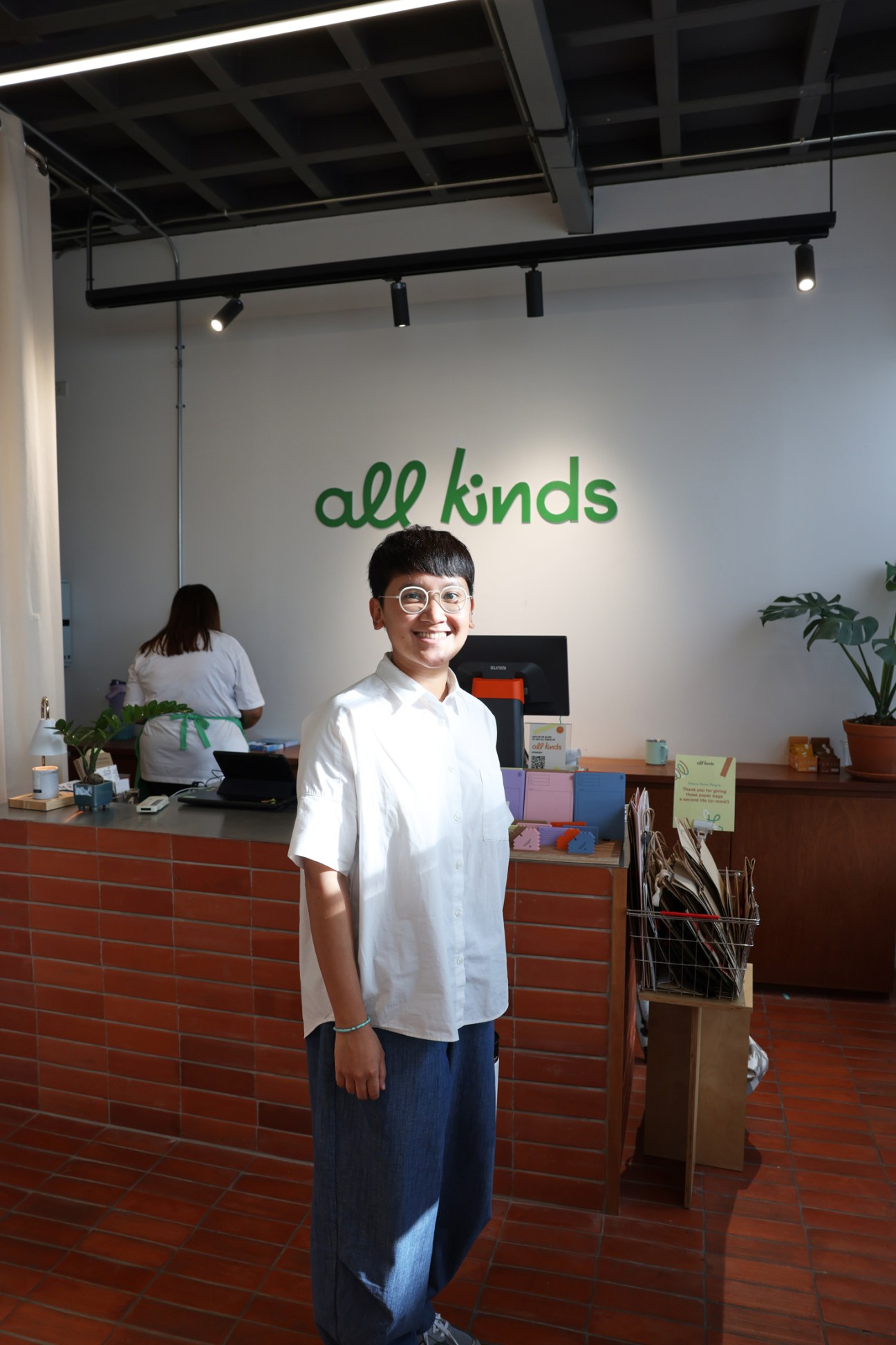Young Asian man in white shirt and glasses standing behind brick counter in modern cafe with 'all kinds' text on wall