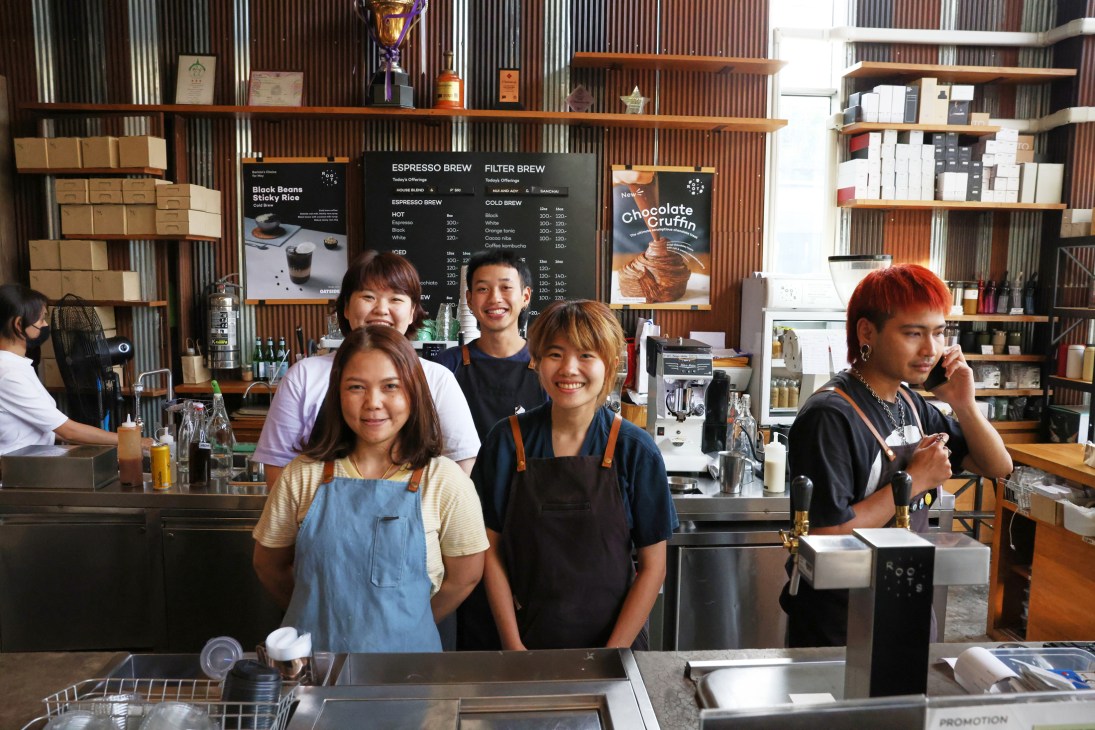 Five smiling staff members behind the counter of a coffee shop with menu boards and shelving visible