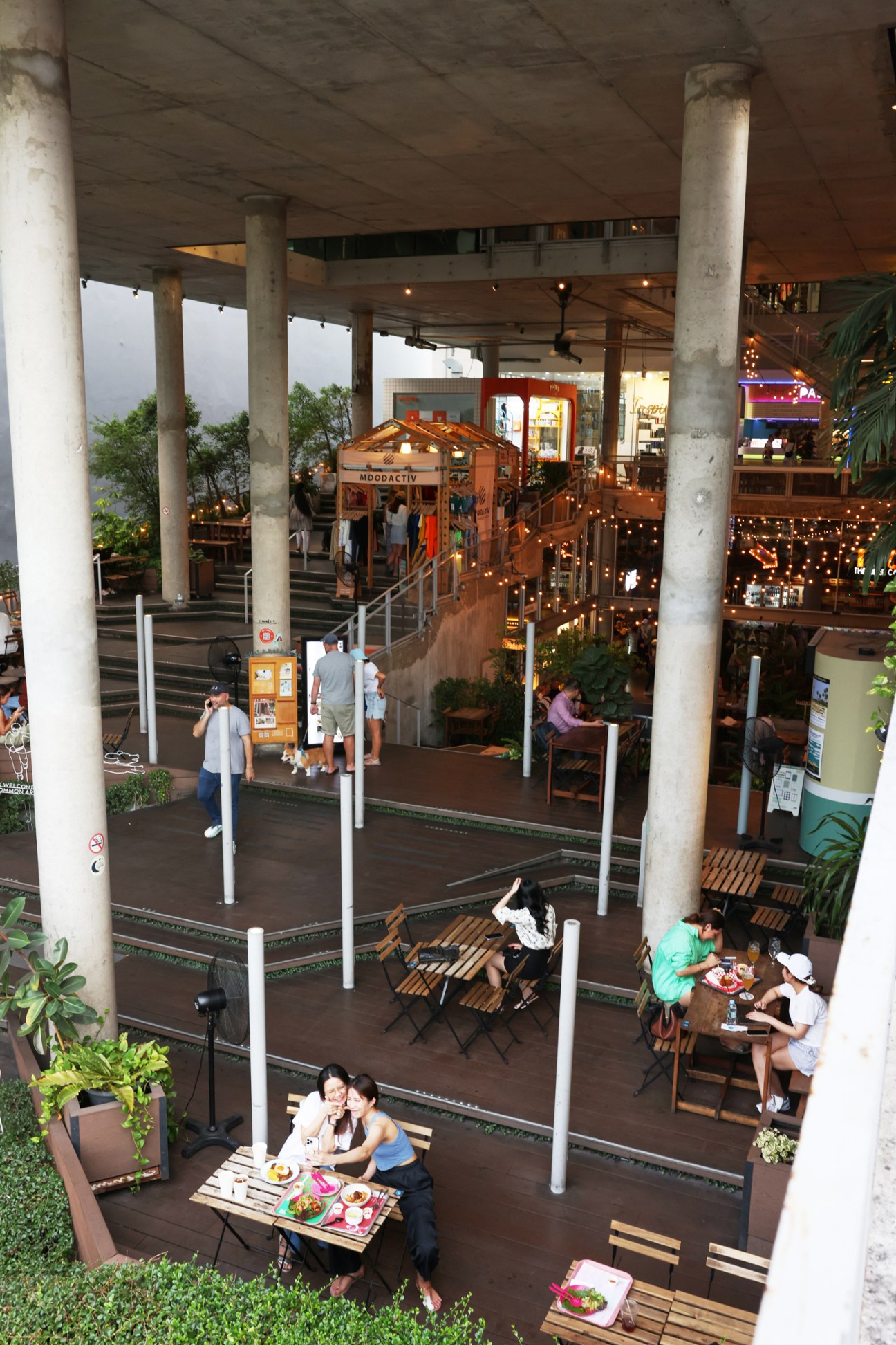 Multi-level food hall with concrete columns, wooden tables, diners eating, and string lights