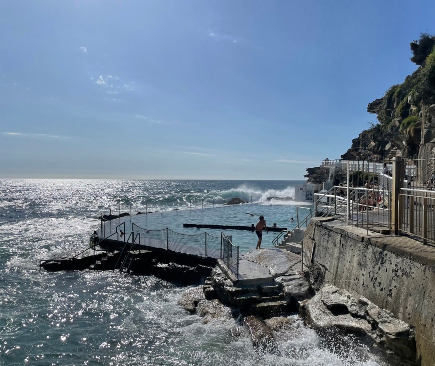 Ocean pool carved into rocky coastline with concrete barriers and metal railings separating it from the sea