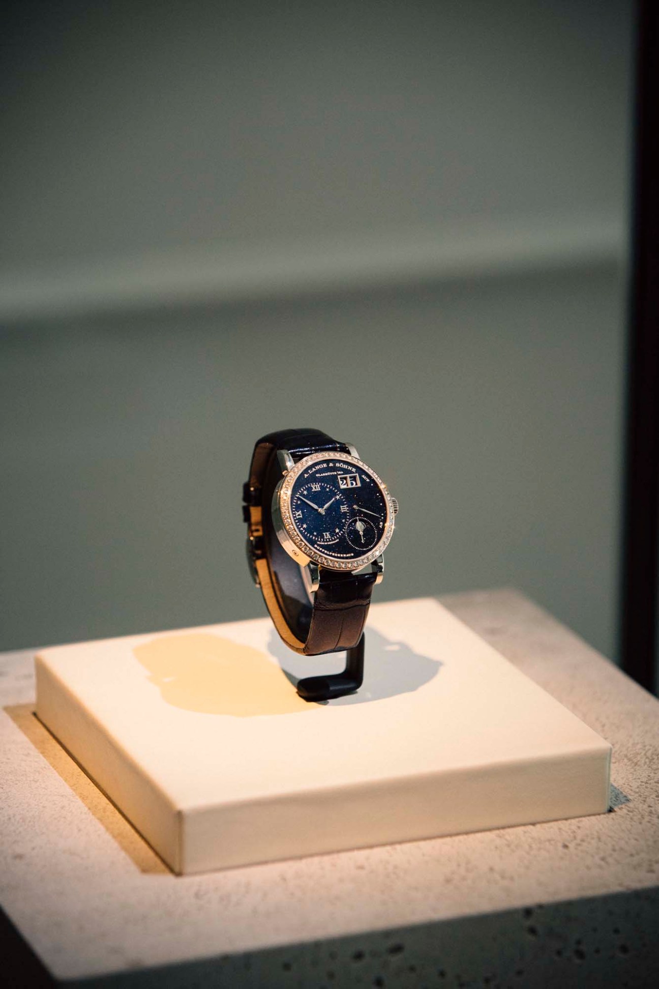 A Lange & Söhne luxury watch with dark blue dial and black leather strap displayed on white platform