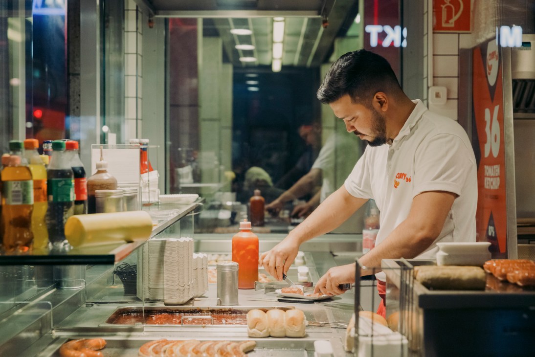 Worker in white uniform preparing currywurst at illuminated food stand counter at night