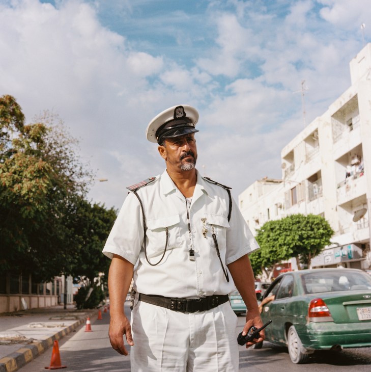 Traffic policeman in white uniform and cap standing on street in downtown Tripoli with cars and buildings behind