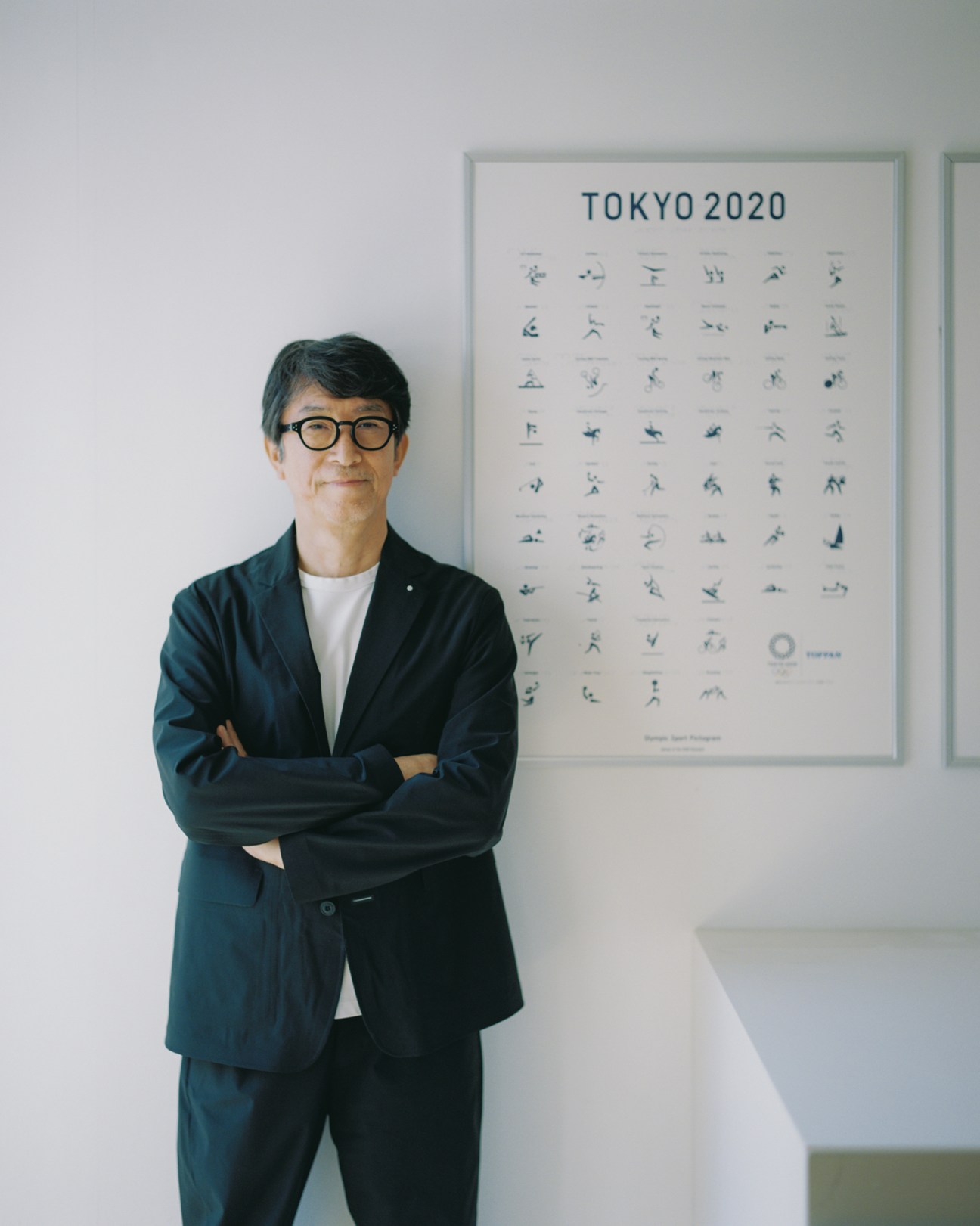 Masaaki Hiromura standing beside a framed poster displaying the complete Tokyo 2020 Olympic pictogram system he designed