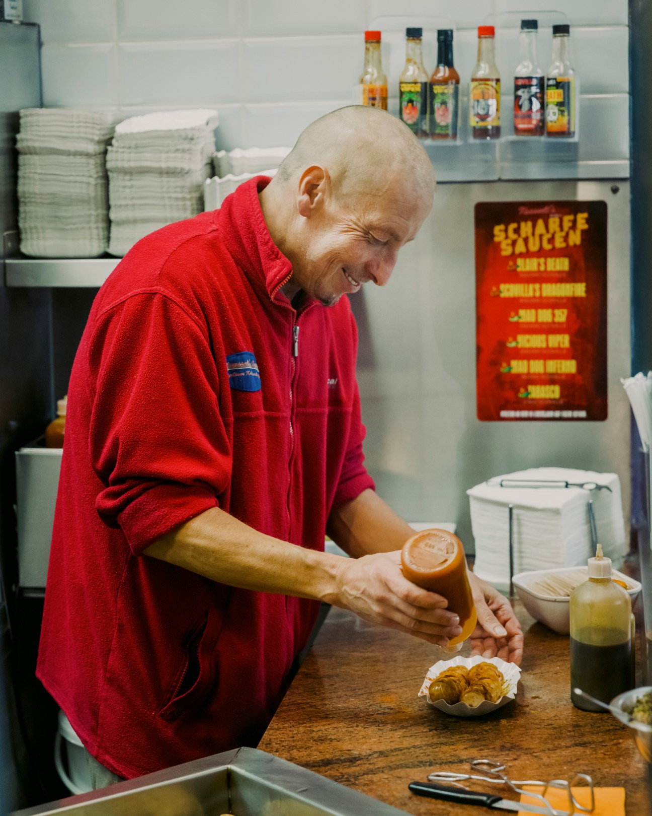 Chef in red jacket pouring curry sauce from bottle onto sliced sausage in commercial kitchen