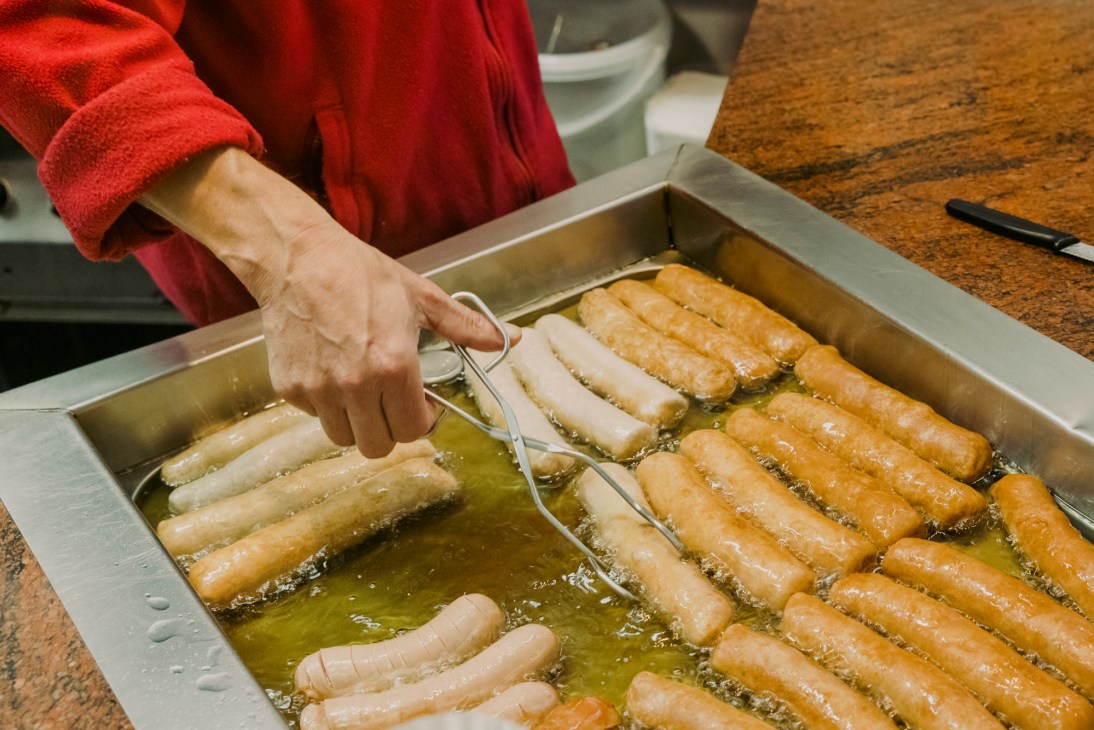 Hand using tongs to turn sausages cooking in bubbling oil in metal cooking trays
