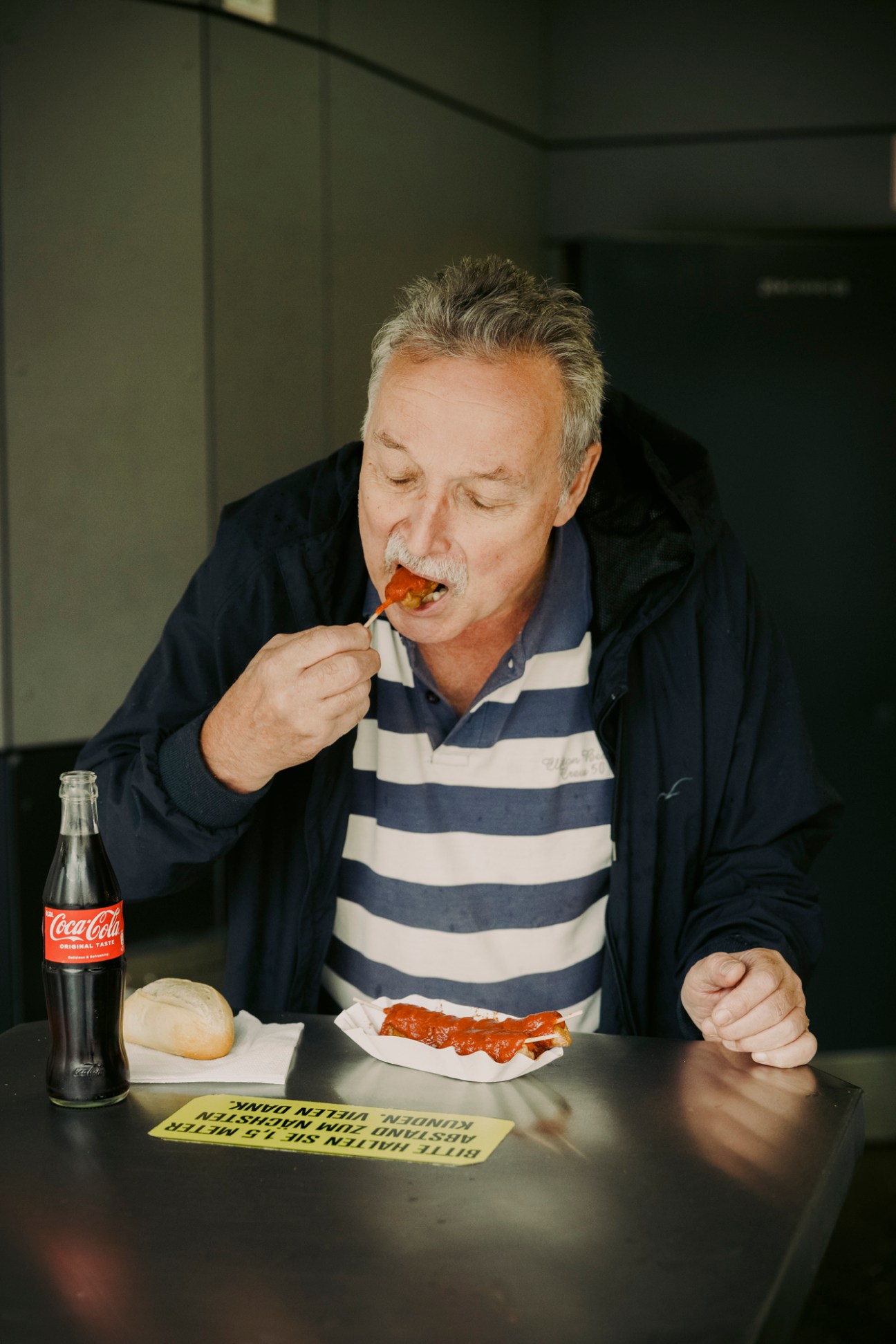 Middle-aged man with gray hair and mustache eating currywurst with wooden fork at table with Coca-Cola bottle
