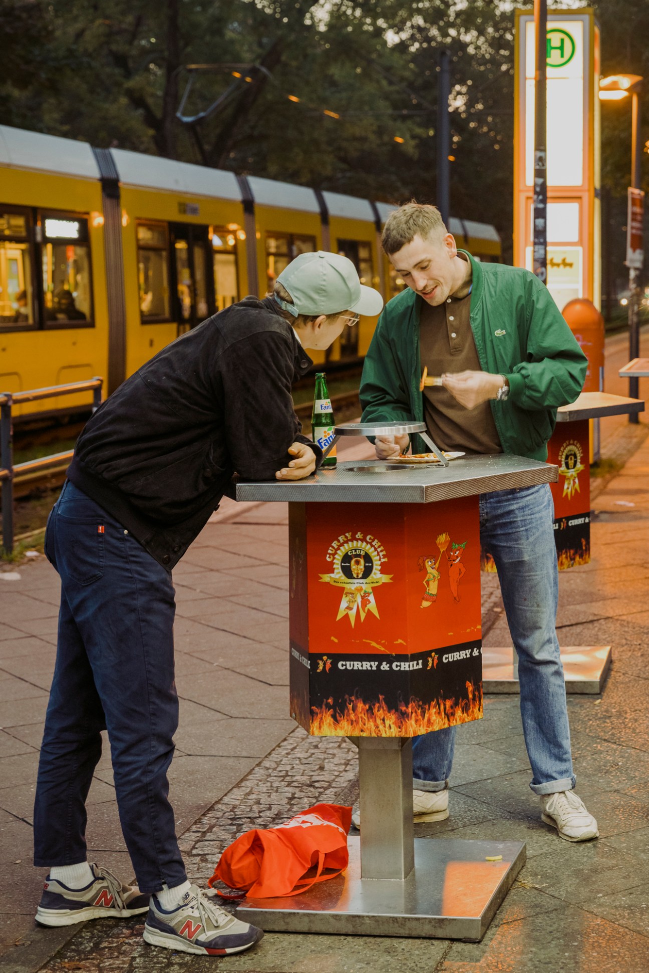 Two people sharing currywurst at street food stand with yellow tram in background