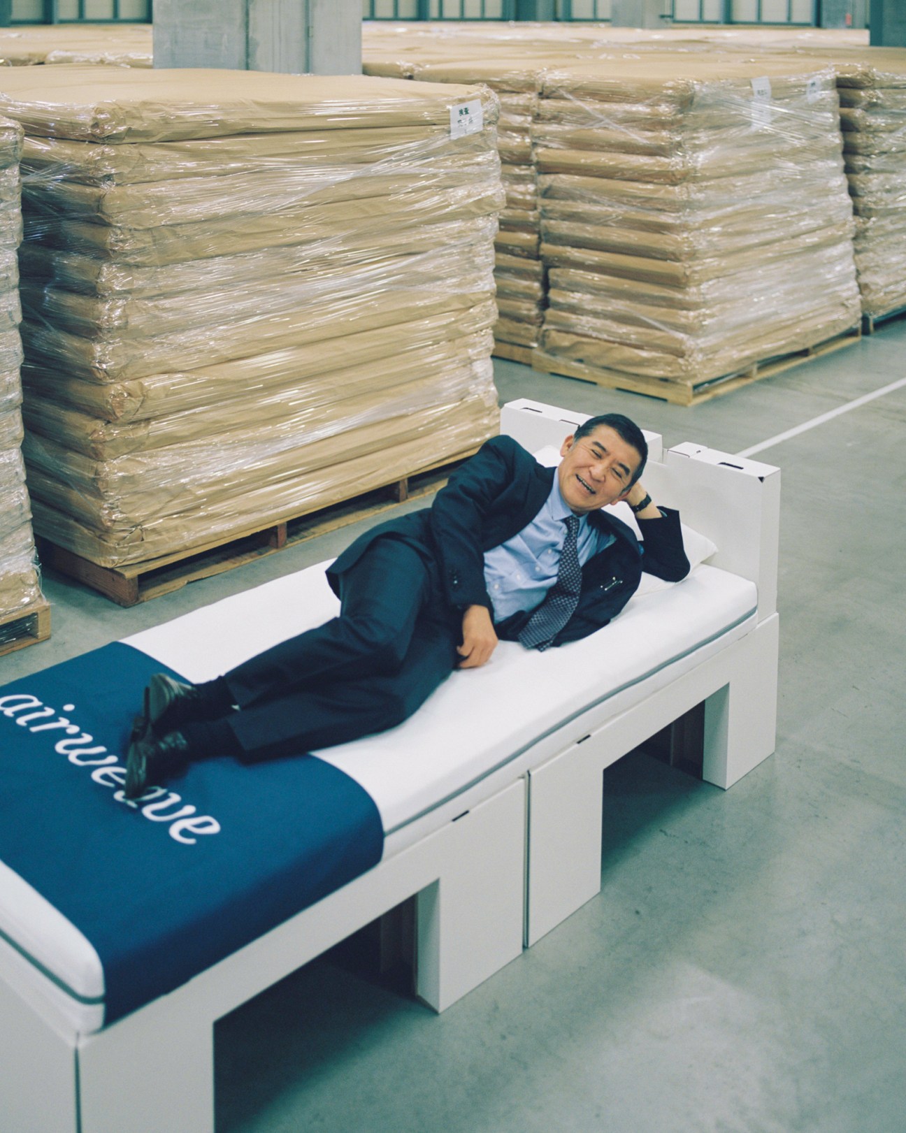 Motokuni Takaoka lying on an Airweave mattress in a business suit in a warehouse setting