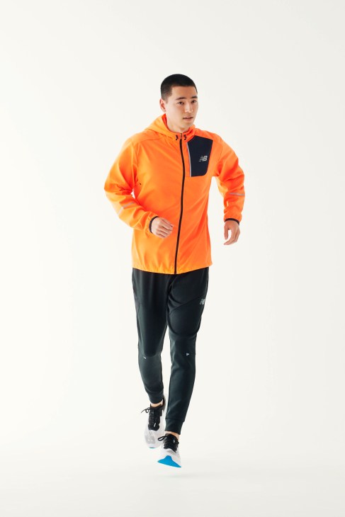 Man jogging in orange New Balance jacket, black trousers, and black trainers against white background.