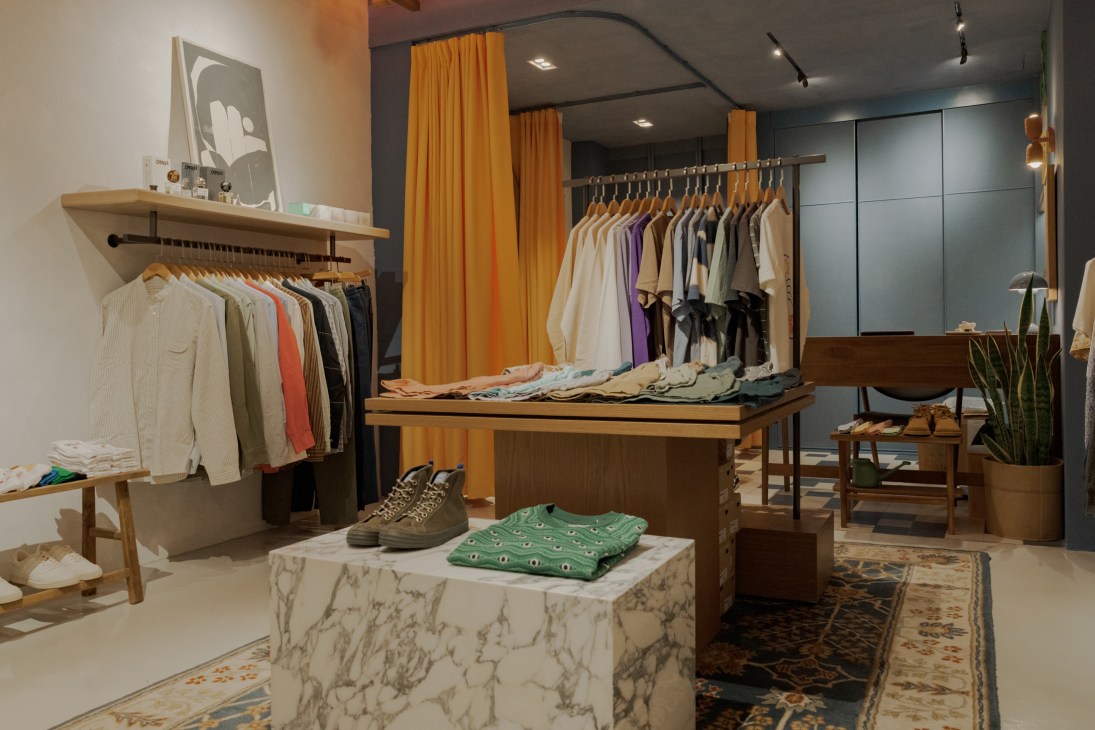 Interior of a clothing retail store with wooden display tables, hanging garments, and orange curtains.
