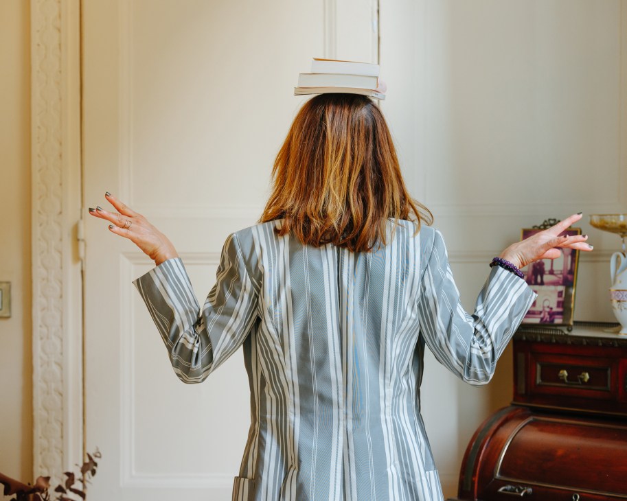 Woman balancing books on her head with arms outstretched in an elegant interior setting