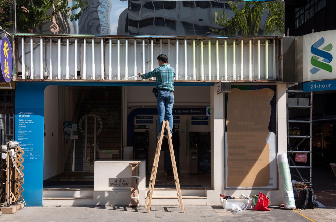 A worker on a wooden ladder adjusts white corrugated metal shutters on a storefront undergoing renovation in Hong Kong.