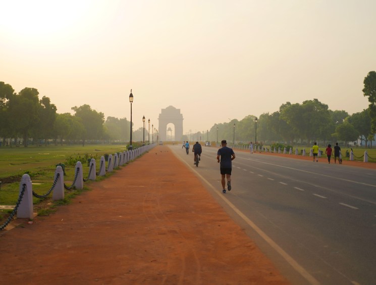 Wide boulevard in New Delhi with joggers and cyclists at dawn, India Gate monument visible in distance