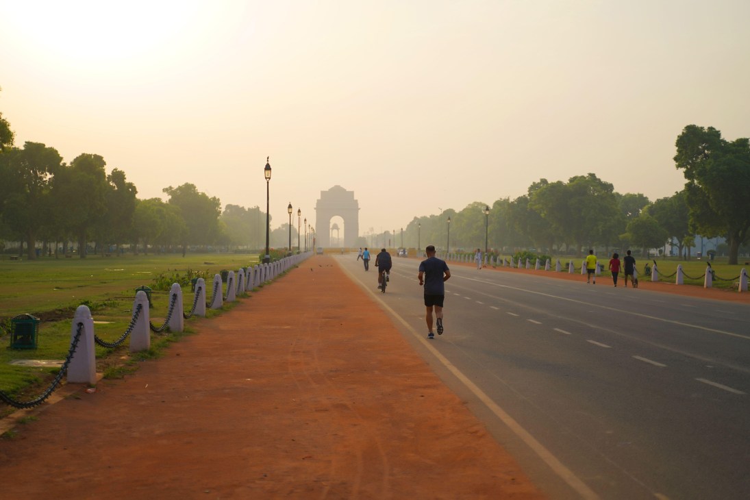 Wide boulevard in New Delhi with joggers and cyclists at dawn, India Gate monument visible in distance