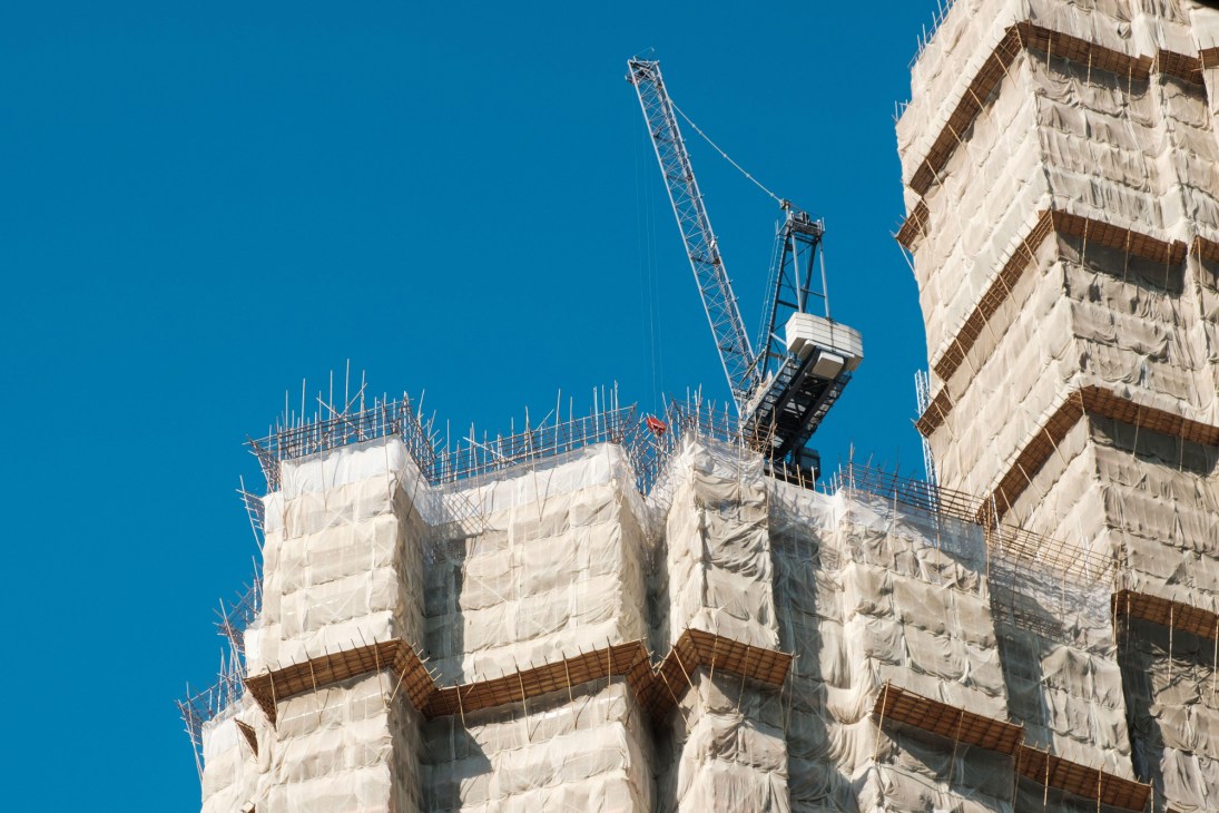 Construction crane hoisting materials above a partially built white concrete high-rise with exposed rebar and safety netting against clear blue sky.