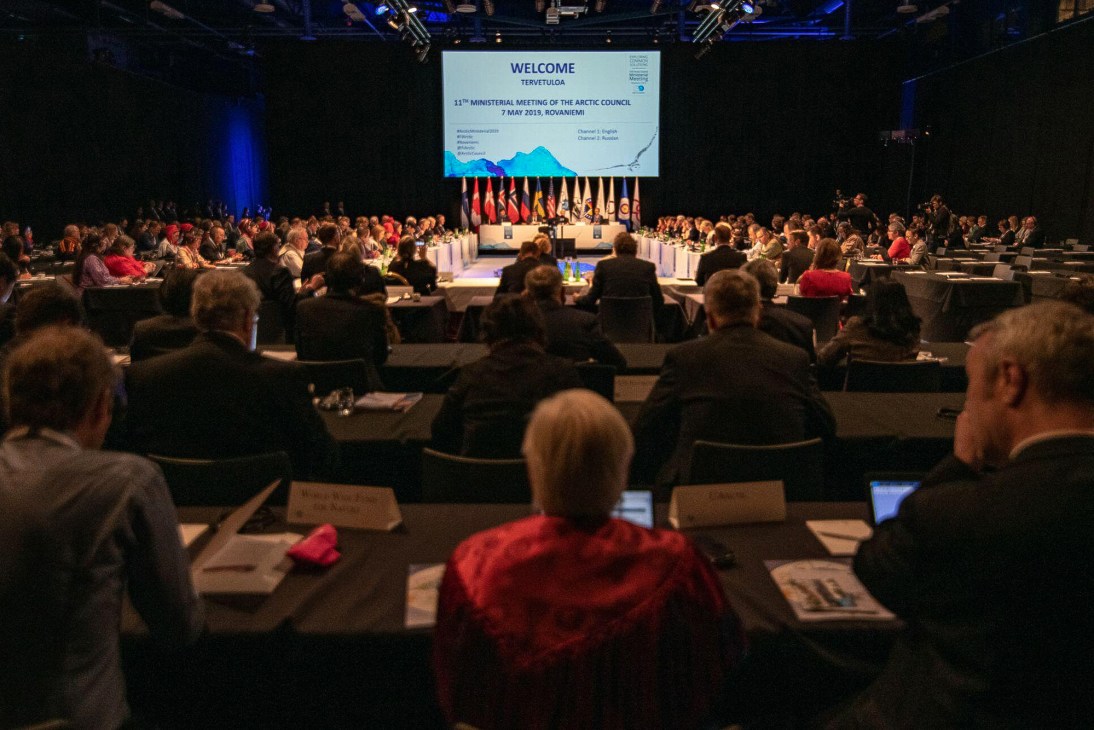 Large conference room with attendees seated at tables facing a stage with welcome screen for Arctic Council meeting