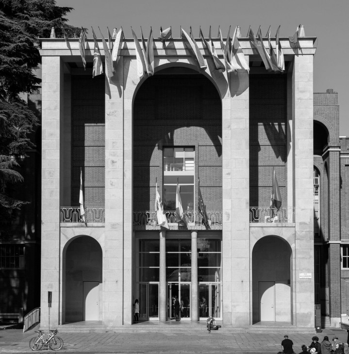 Modernist building facade with large arches, classical columns, and flags displayed on upper level balcony