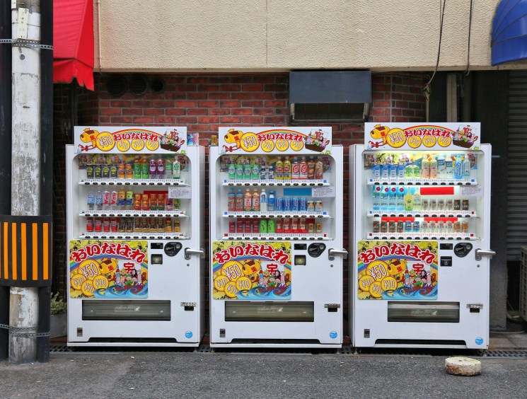 Three Japanese vending machines displaying colorful drinks and snacks lined up on a street