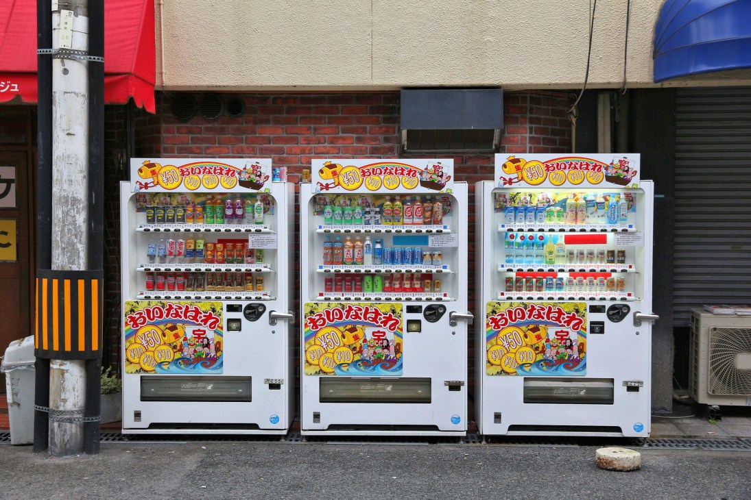 Three Japanese vending machines displaying colorful drinks and snacks lined up on a street