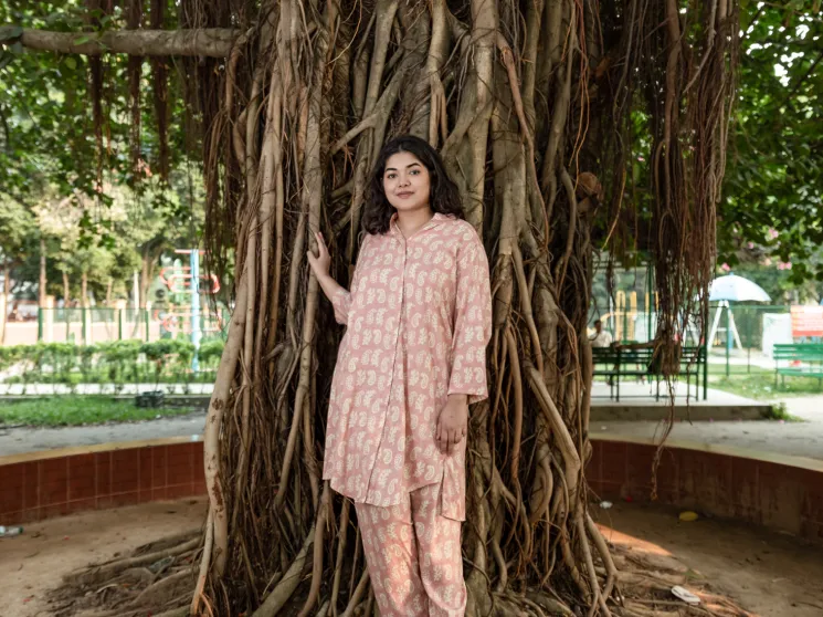 Bushra Afreen stands against large tree roots in pink patterned outfit in urban park setting