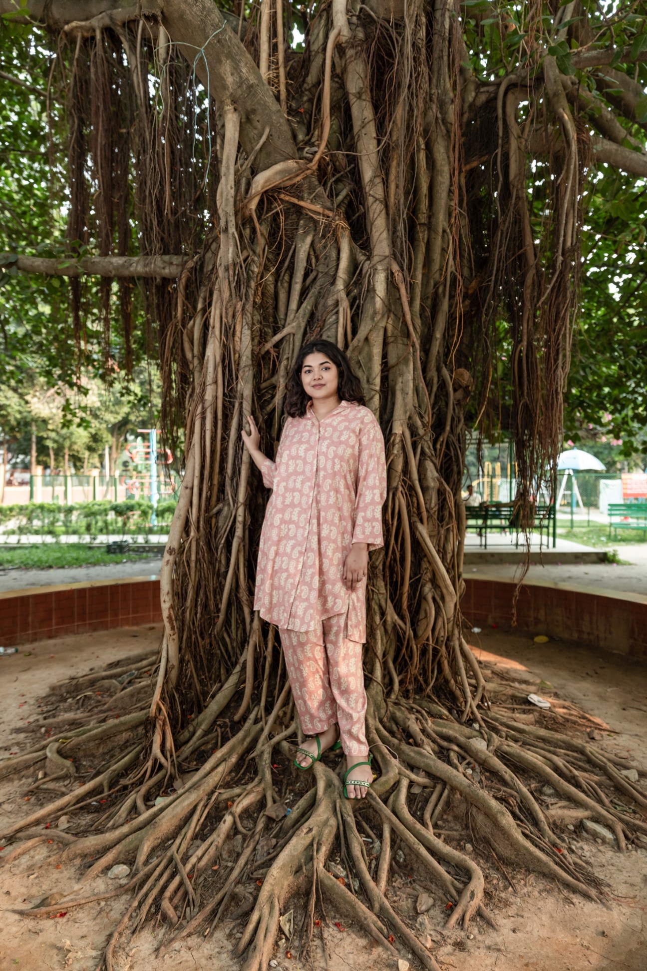 Bushra Afreen stands against large tree roots in pink patterned outfit in urban park setting