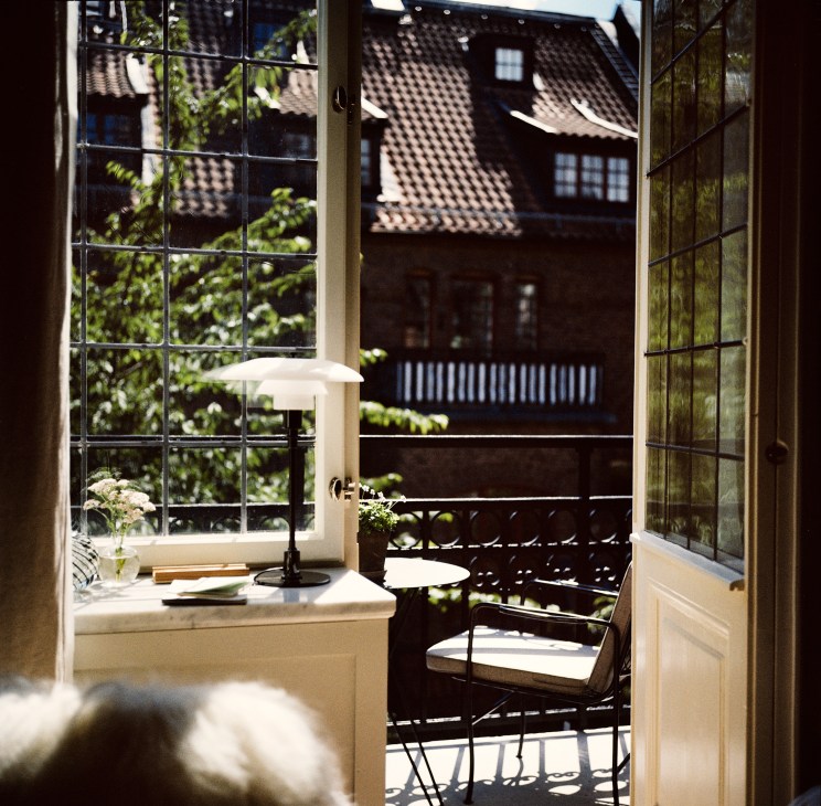 View through open balcony doors showing desk area with lamp and chair overlooking courtyard with traditional tiled roofs