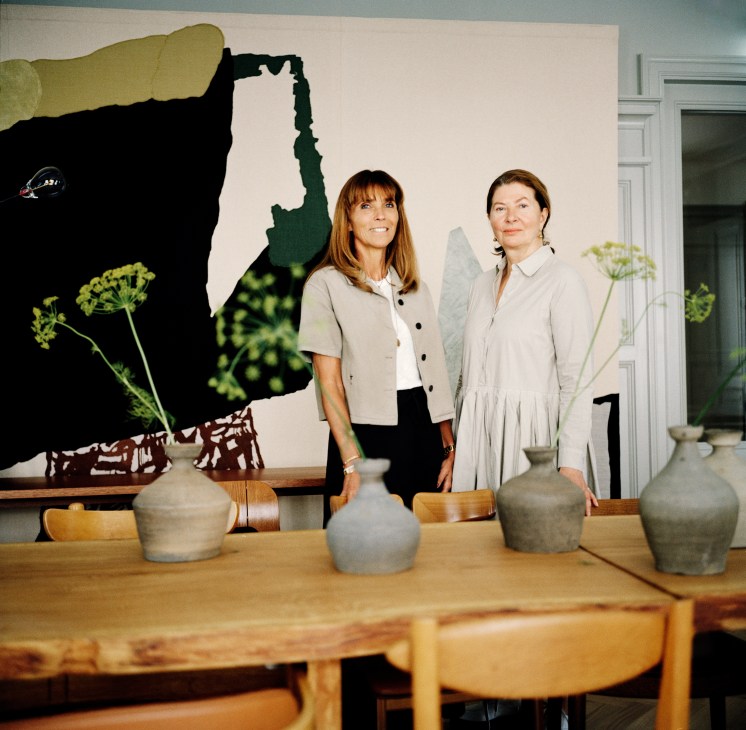 Jeanette Mix and Ilse Crawford standing at a wooden table with pottery vessels and green botanical elements, with abstract artwork on wall behind.