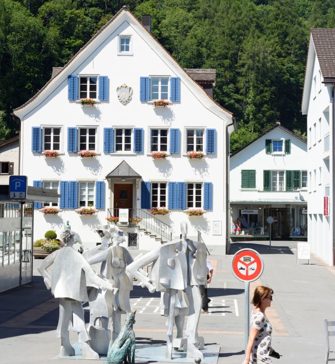 White-walled historic building with blue shutters and flower boxes in St Gallen, Switzerland's town center surrounded by green forest.
