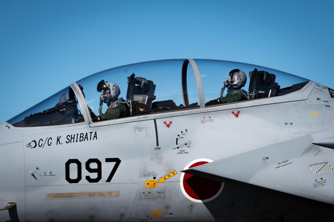 Two pilots in helmets sit in cockpit of Japanese military aircraft numbered 097 with name C/C K. SHIBATA visible