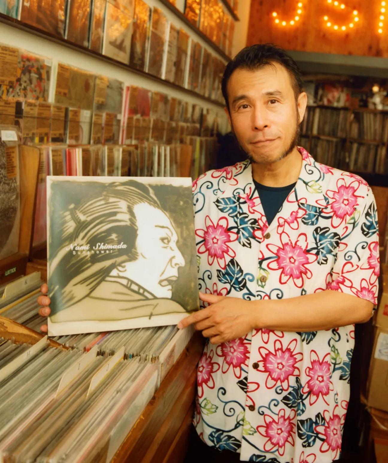 Soichi Terada holds a vinyl record in a record shop surrounded by shelves of music albums.