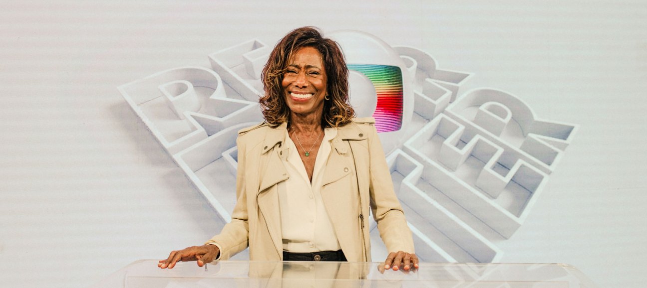 Glória Maria sitting at a broadcast desk in a television studio