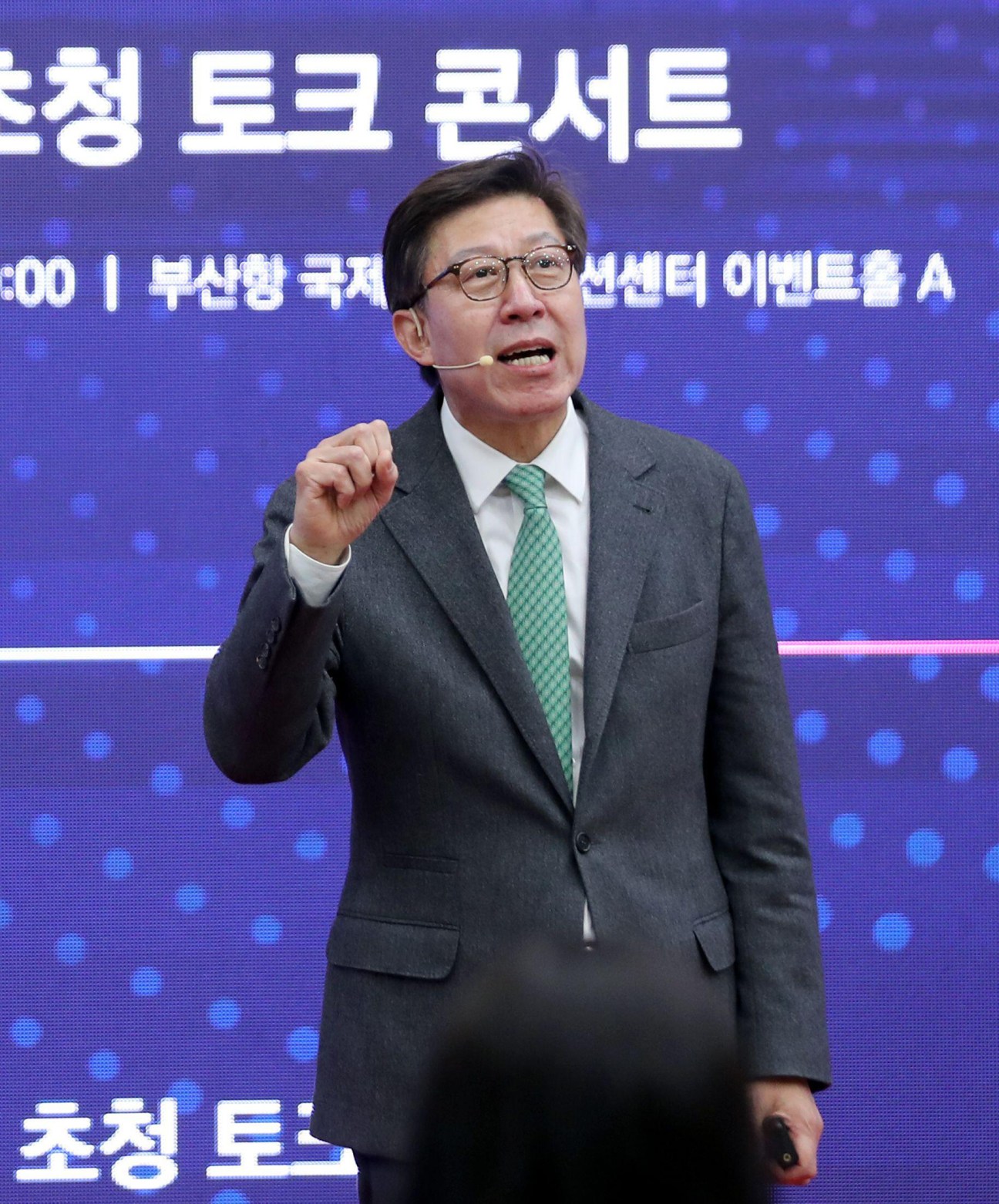 Man in dark suit and green tie speaking at podium with Korean text on blue background