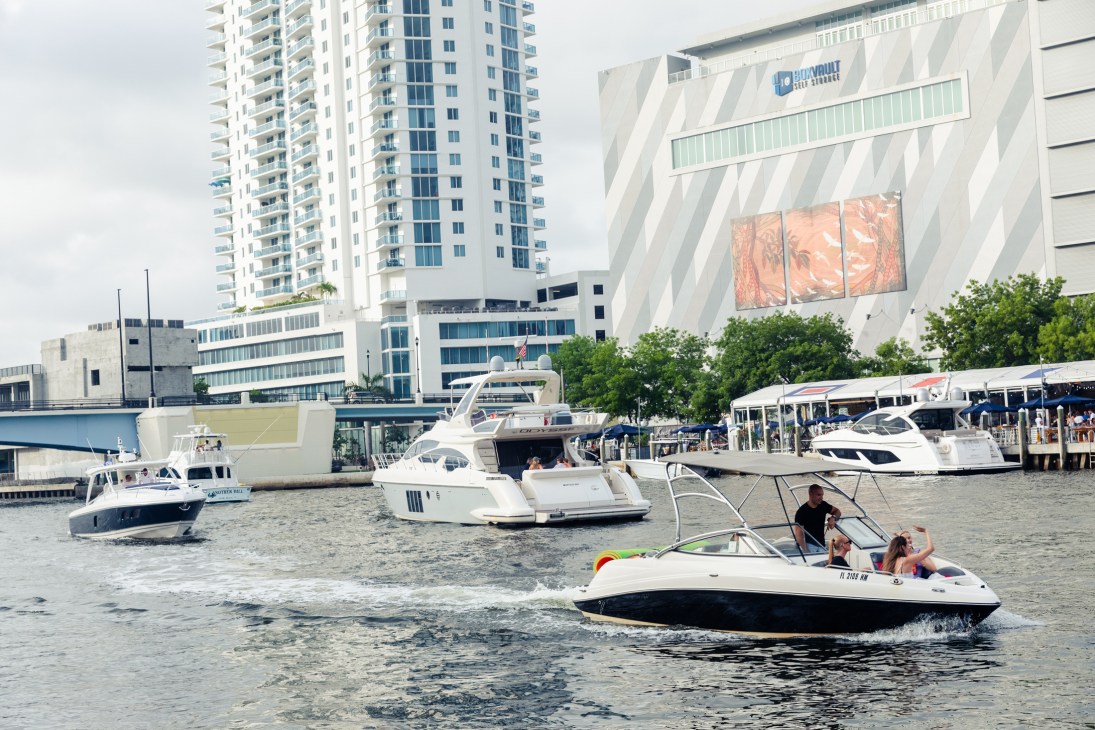 Multiple boats on water with modern buildings and marina in background