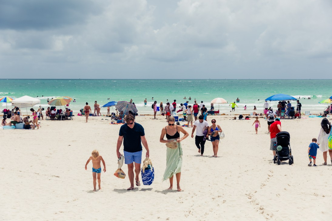 Families with beach umbrellas and children walk on crowded white sand beach with turquoise ocean