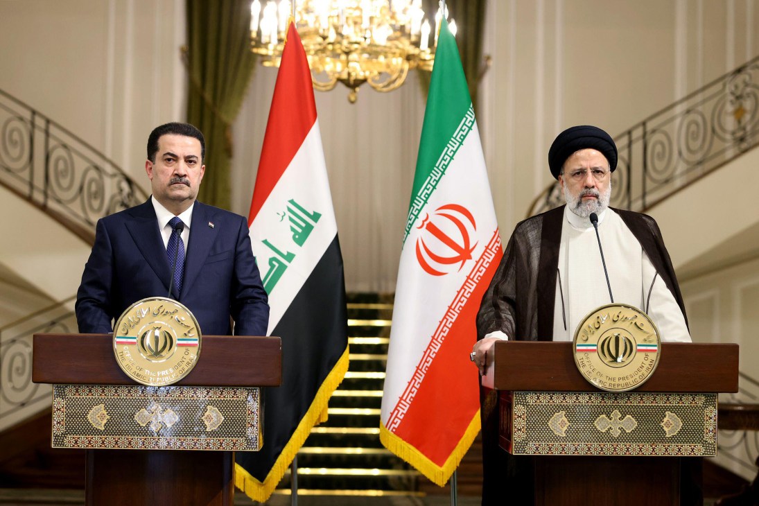 Two officials at podiums with Iraqi and Iranian flags during diplomatic meeting
