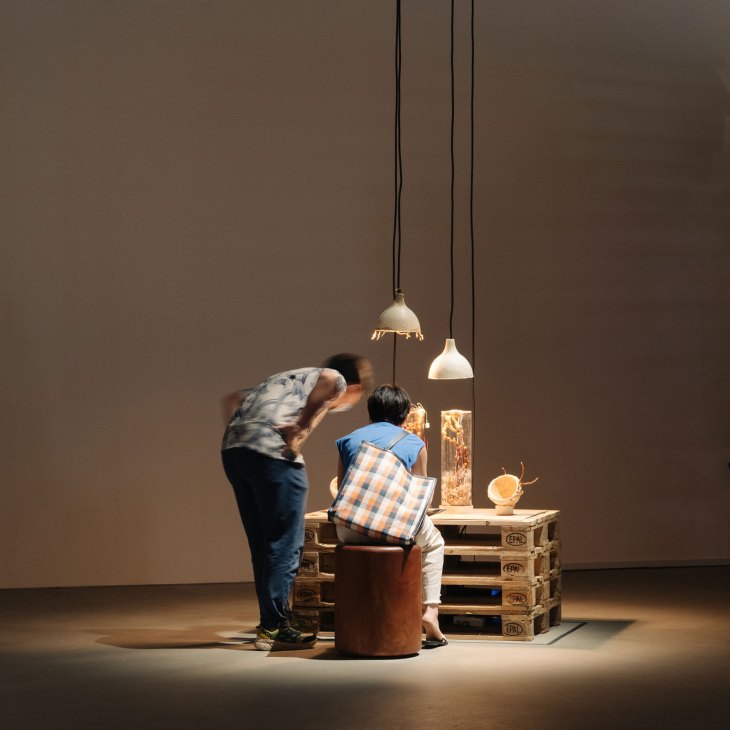 Two visitors examine an art installation with pendant lights and objects on wooden pallets in a gallery space.