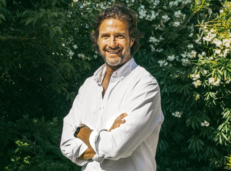 A smiling middle-aged man in a white shirt and jeans stands with arms crossed in a lush garden.