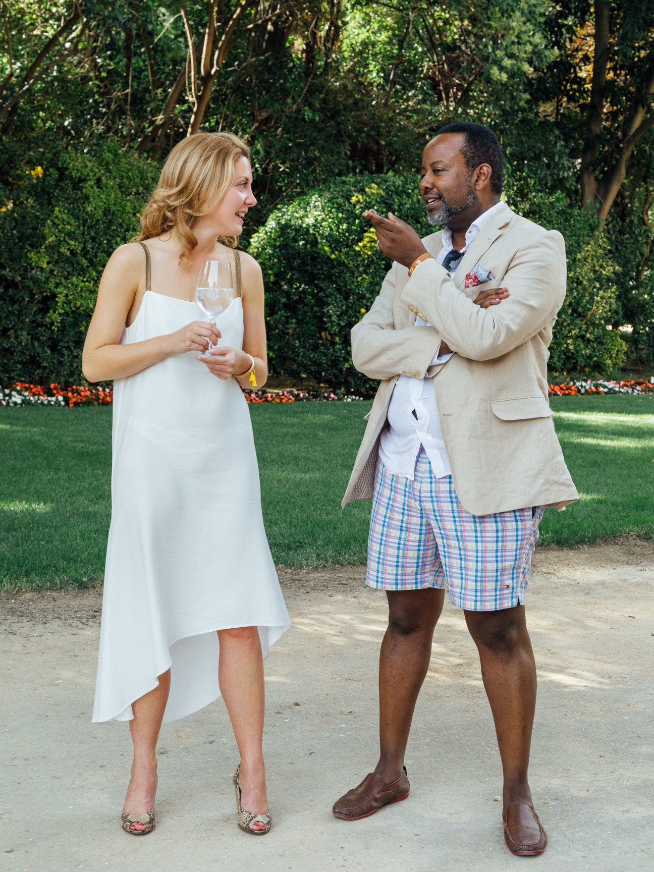 Two people conversing at an outdoor event, woman in white dress holding wine glass, man in beige blazer