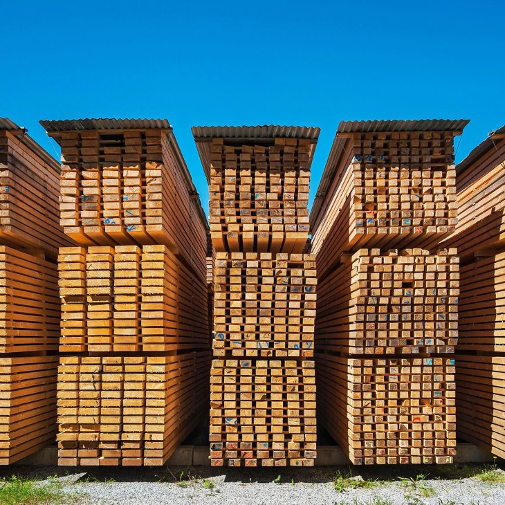 Stacked wooden lumber and timber planks piled high against a clear blue sky.
