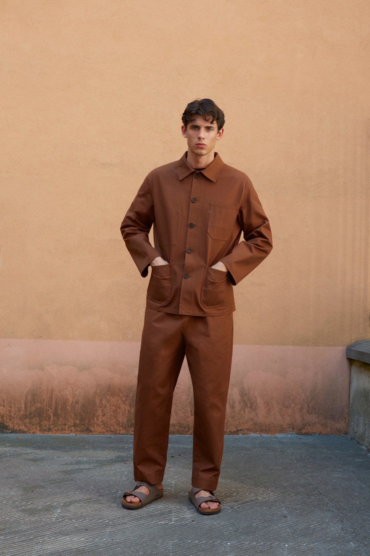 Man in brown matching jacket and trousers with hands in pockets standing against beige wall