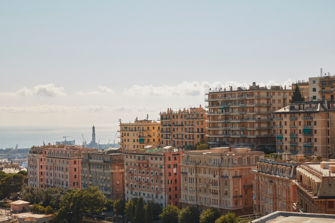 Aerial view of Italian city buildings with warm-toned architecture and coastal backdrop under cloudy sky